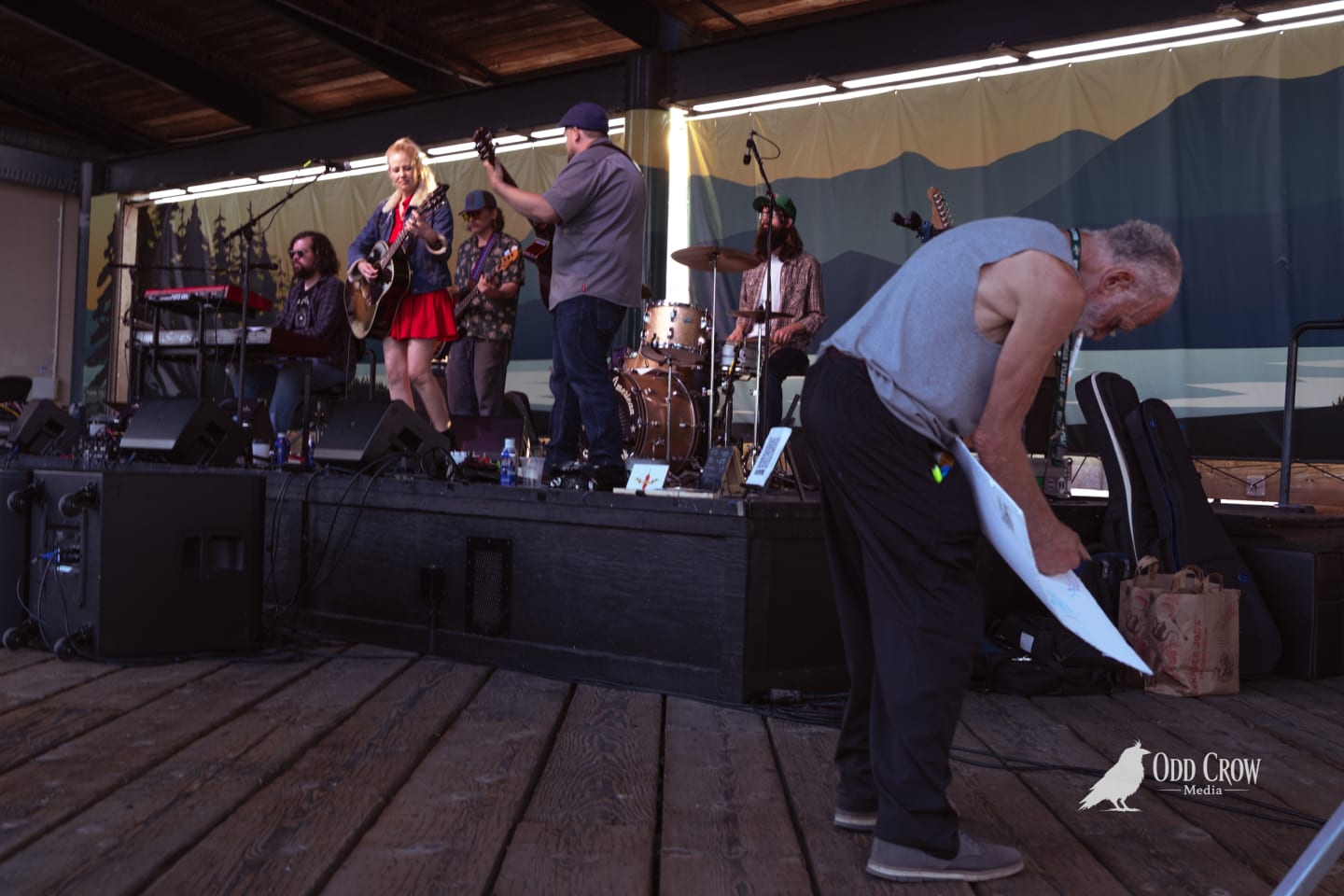 Band performs on outdoor stage while older man bends down in foreground at waterfront venue.