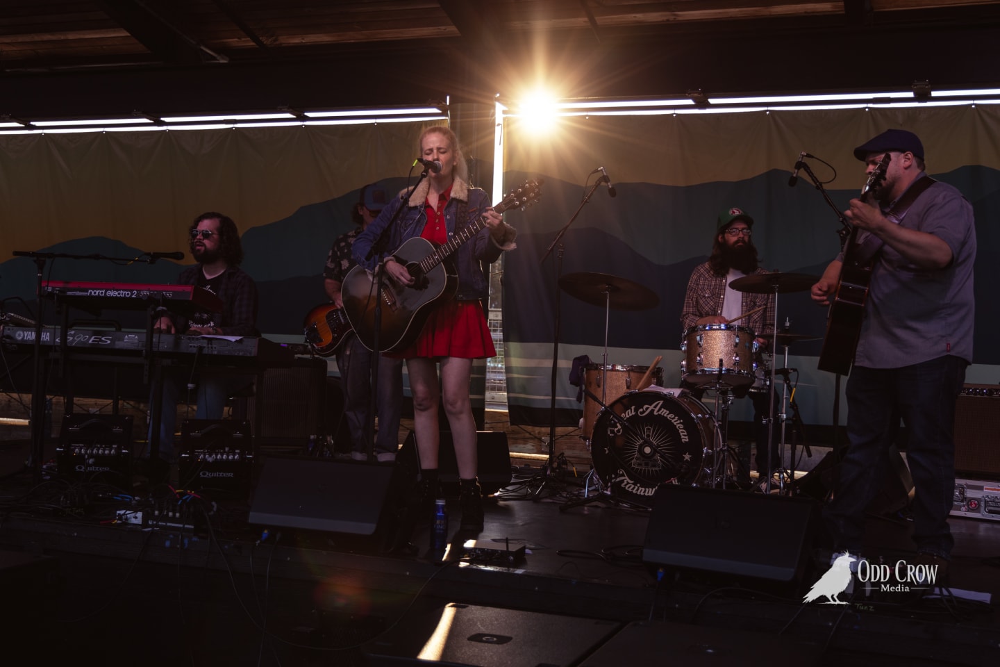 Female vocalist in red dress plays acoustic guitar onstage with her band at an outdoor pier venue.