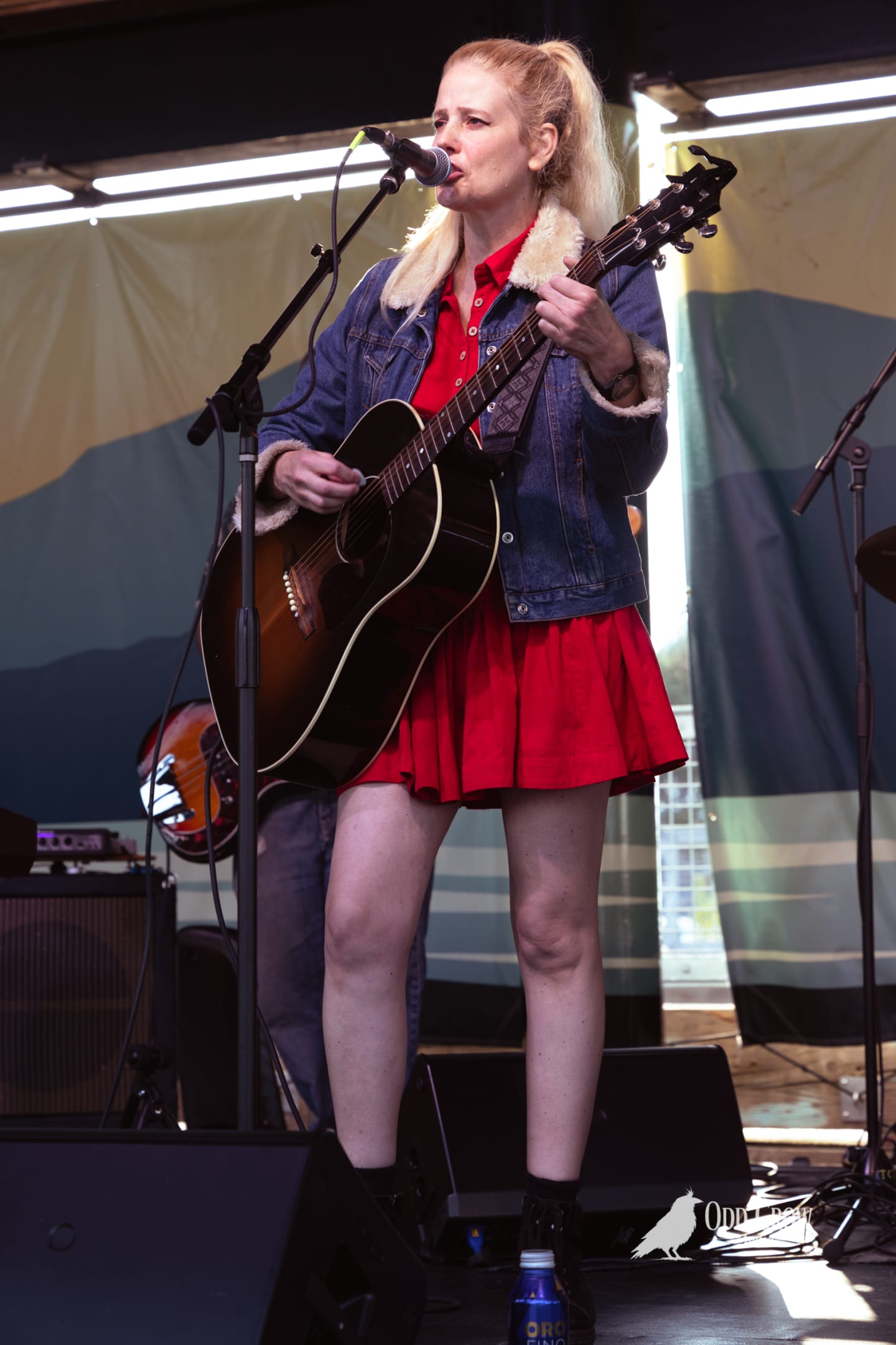 Blonde woman in red dress and denim jacket plays acoustic guitar and sings on outdoor stage.