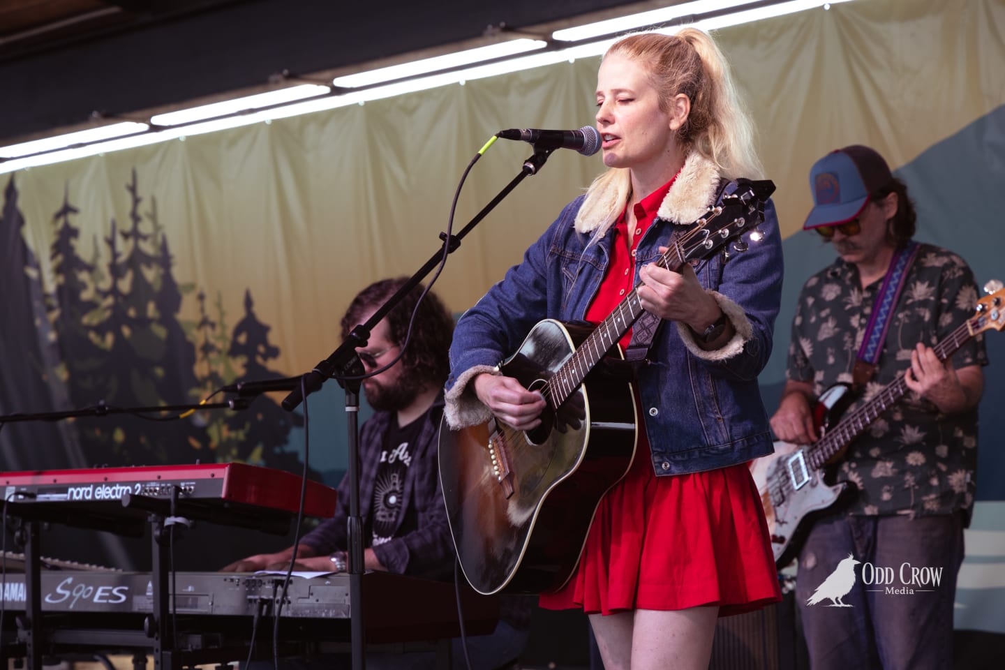 Blonde female vocalist in red dress and denim jacket plays acoustic guitar on outdoor stage with bandmates.