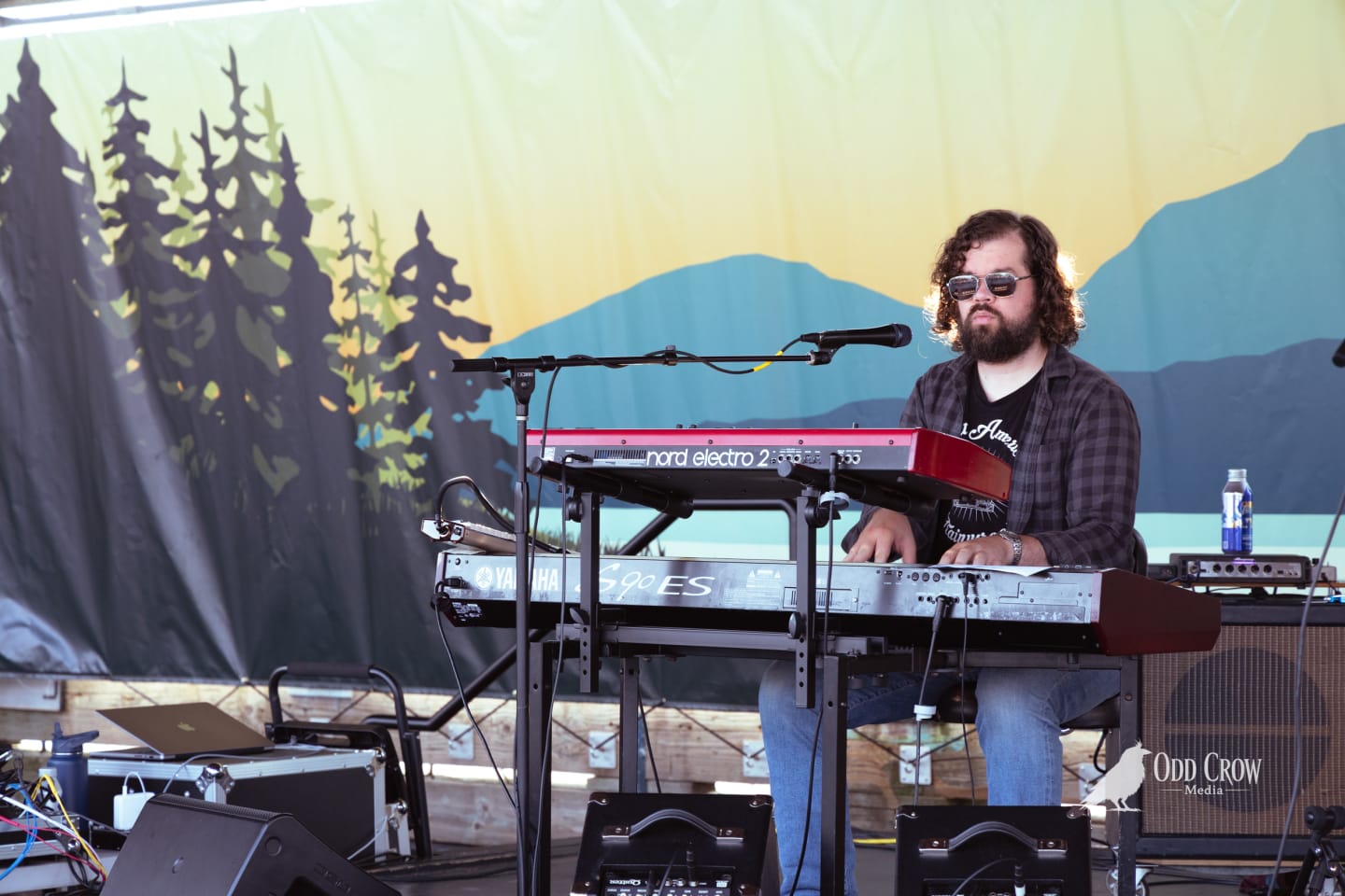 Bearded keyboardist plays Nord Electro 2 and Yamaha S90 ES on an outdoor stage with nature mural backdrop.