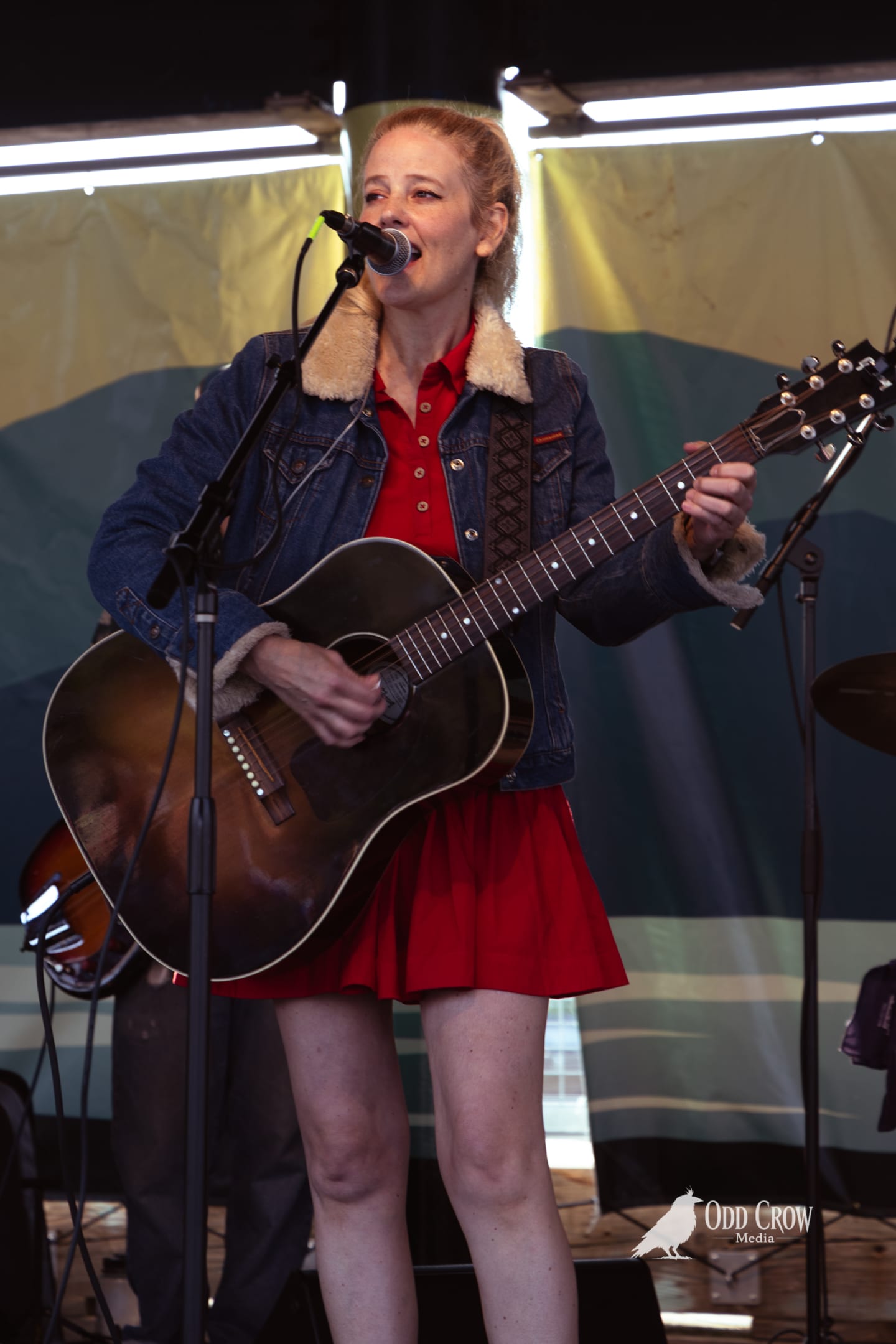 Woman in red dress and denim jacket singing and playing acoustic guitar on outdoor stage.