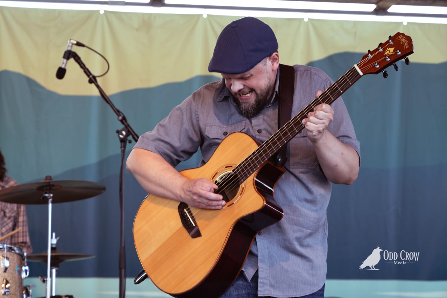 Bearded guitarist in blue cap plays acoustic guitar onstage, smiling, at an outdoor festival performance.
