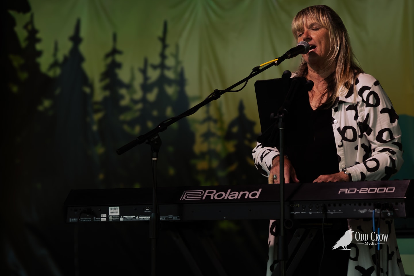 Blonde woman singing into microphone while playing Roland RD-2000 keyboard on stage with forest backdrop.