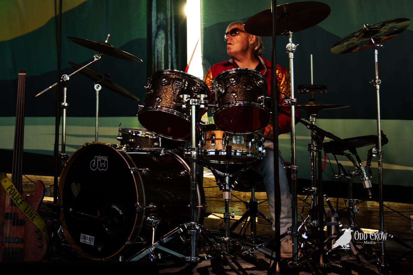 Drummer in red jacket and sunglasses plays DW kit on an outdoor stage.