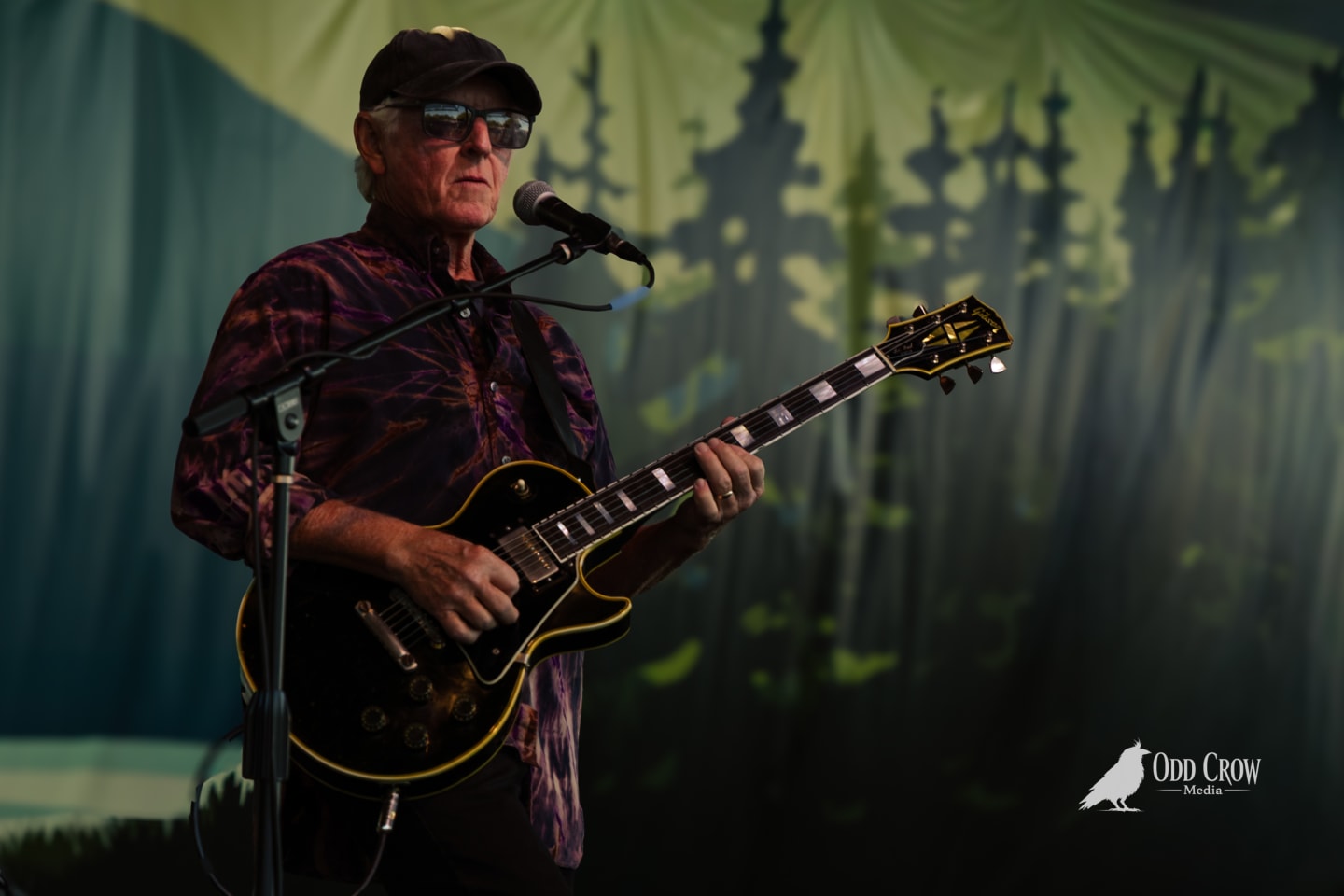 Older guitarist in cap and sunglasses playing a Gibson Les Paul on a decorated outdoor stage.