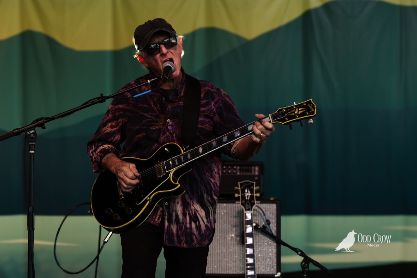 Guitarist in tie-dye shirt and black cap performs on outdoor stage with Gibson Les Paul.