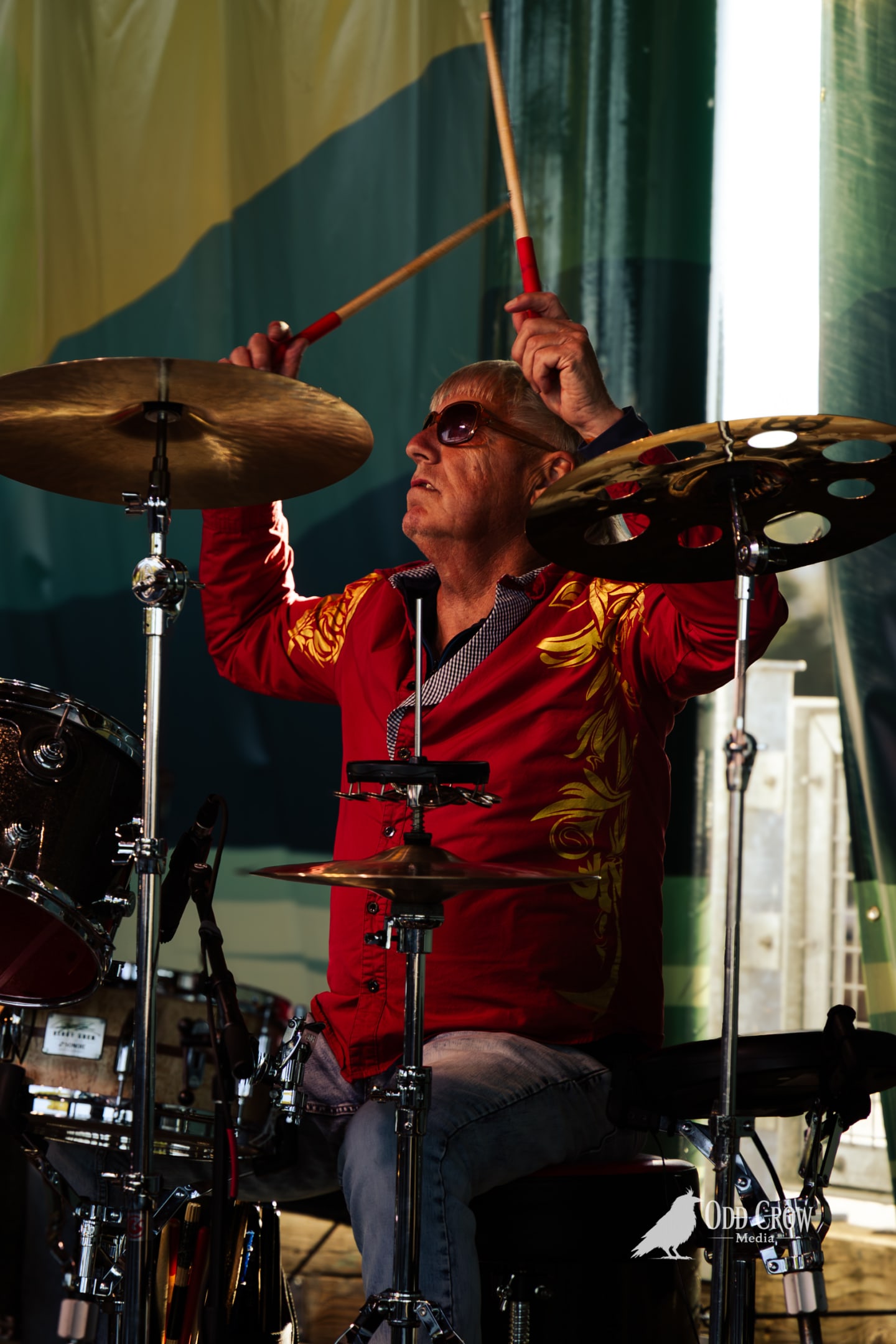 Drummer in red embroidered jacket and sunglasses strikes cymbals mid-performance on an outdoor stage.