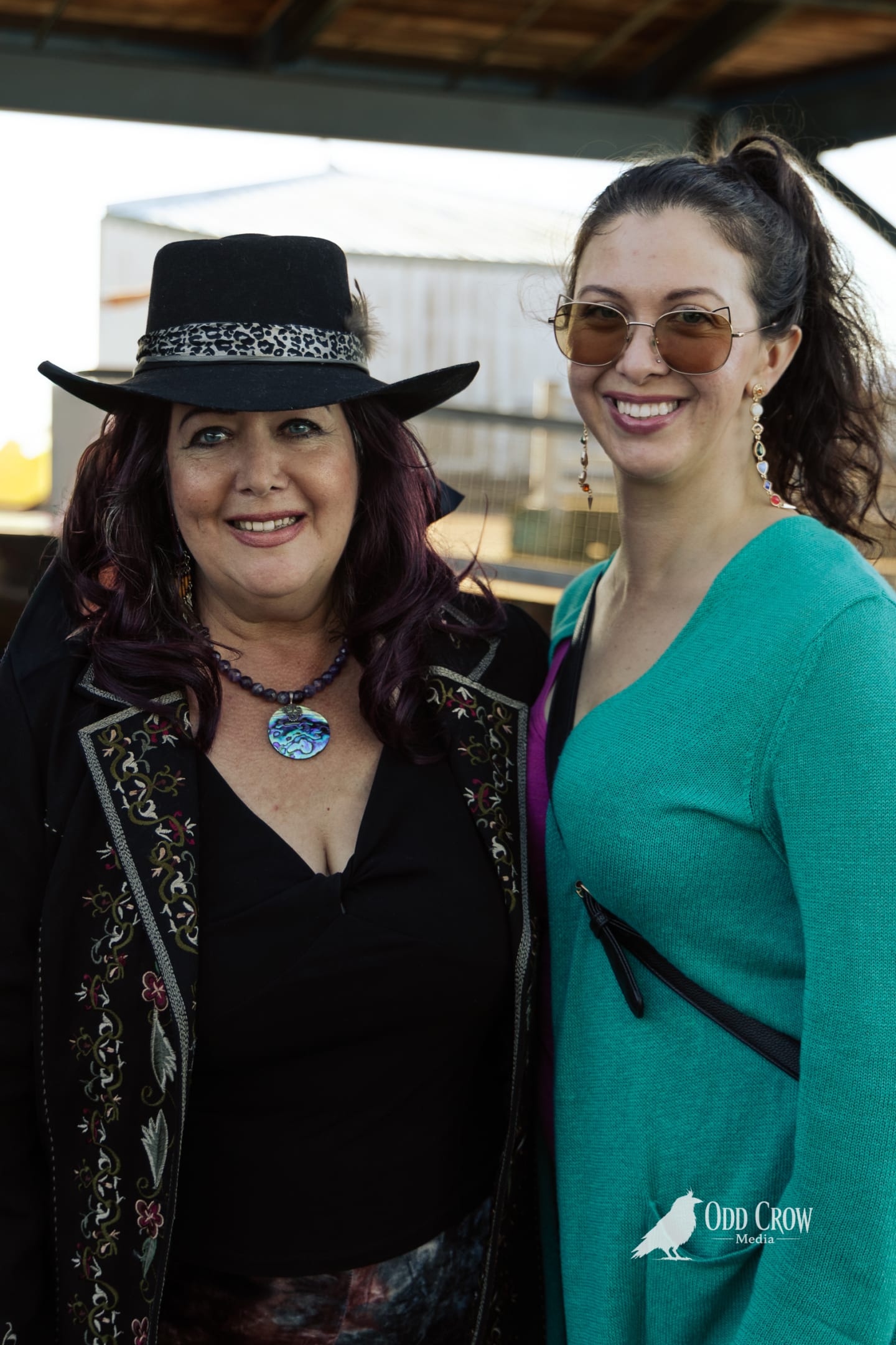 Two women smiling together outdoors at Arts in Action, one wearing a black hat and embroidered coat.