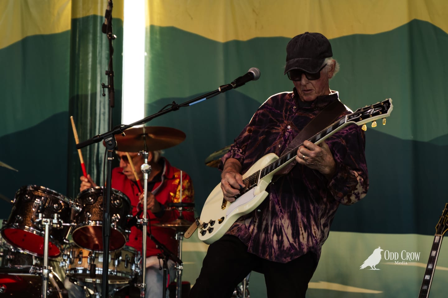 Guitarist in tie-dye shirt and cap plays white electric guitar on outdoor stage, drummer visible behind.