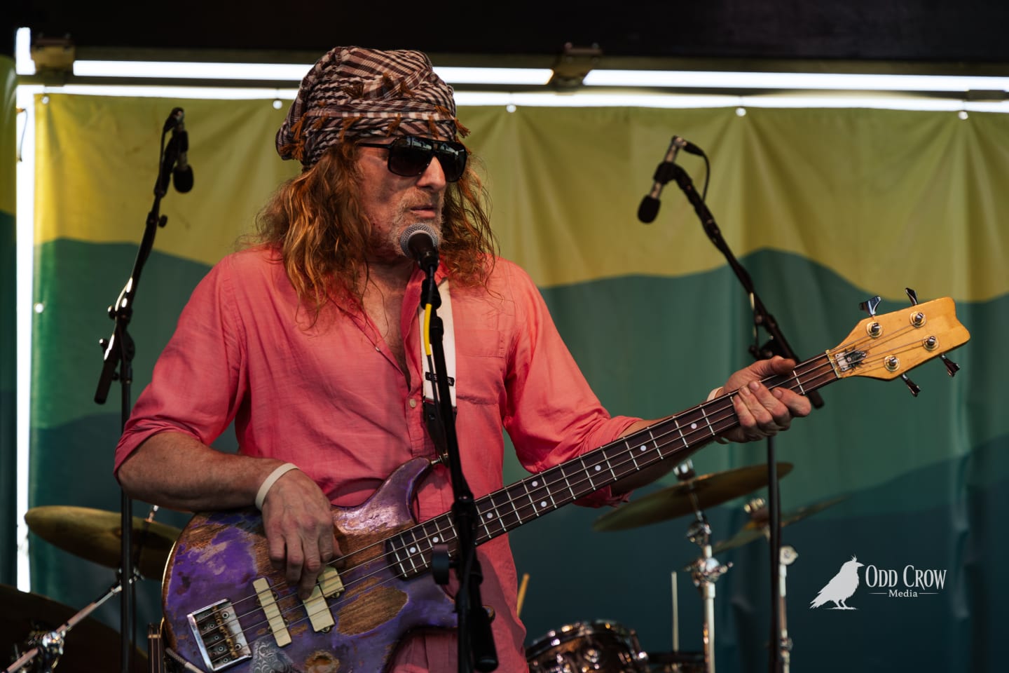Bass guitarist with long red hair, head wrap, and sunglasses performing on stage at an outdoor festival.