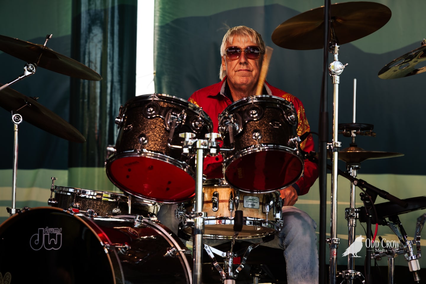 Drummer in red shirt and sunglasses plays DW kit on stage at outdoor concert.