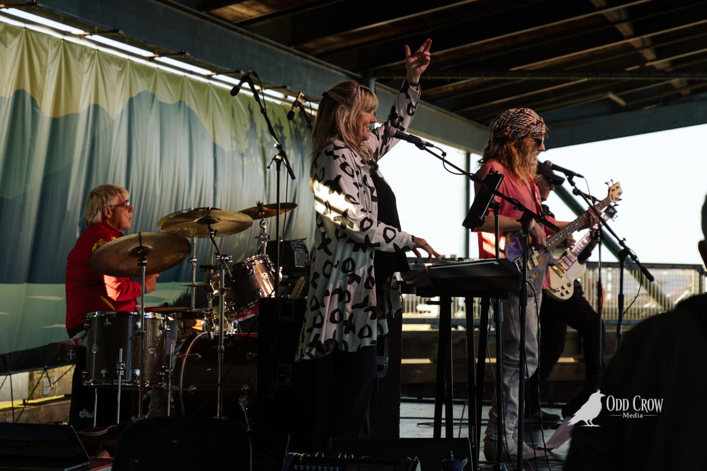 Keyboardist raises her hand mid-performance while bandmates play drums and guitar on an outdoor covered stage.