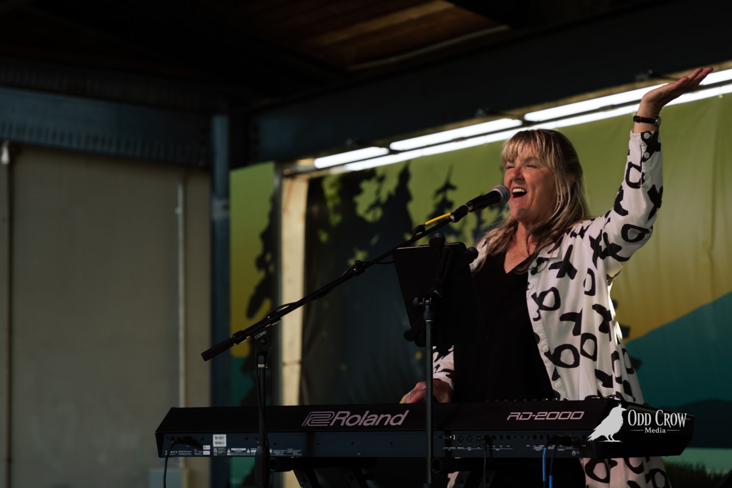 Female keyboardist singing joyfully at Roland RD-2000, one arm raised, performing outdoors at Arts in Action festival.