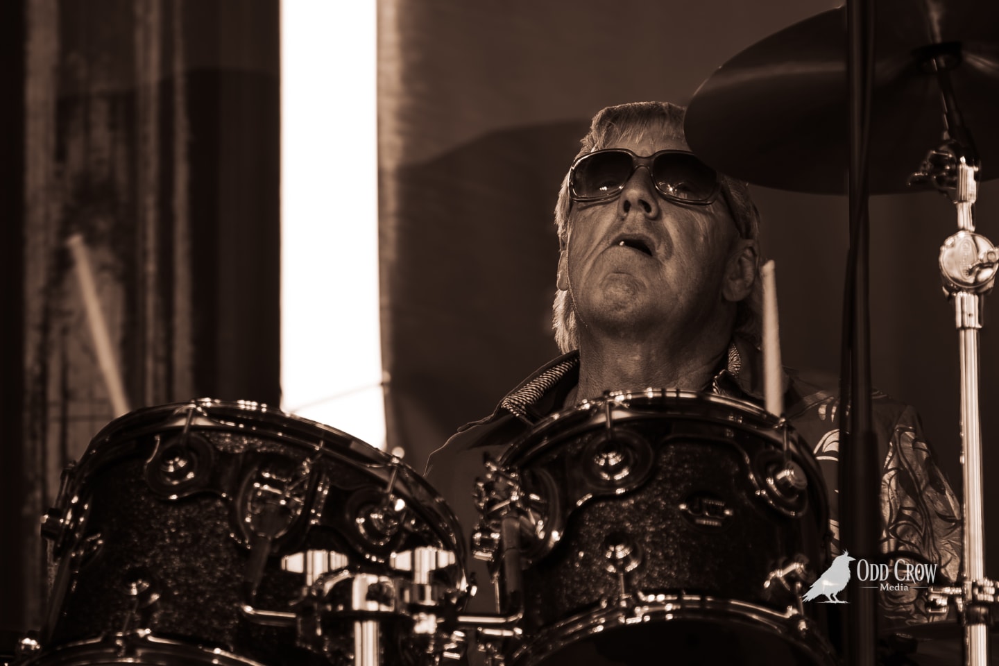 Drummer wearing sunglasses plays ornate drum kit on stage, head tilted back mid-performance.