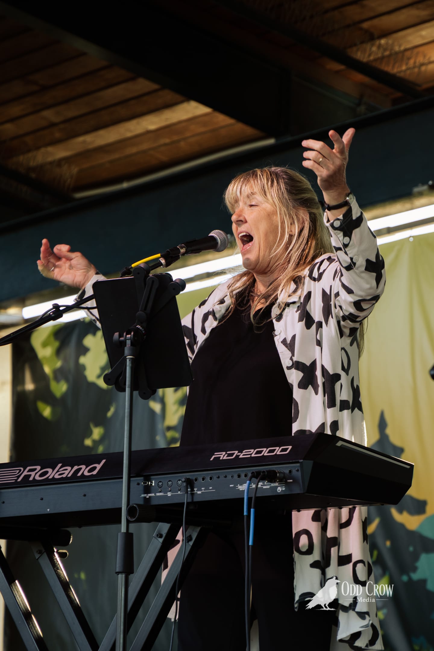Woman singing with arms raised behind a Roland RD-2000 keyboard on an outdoor stage.
