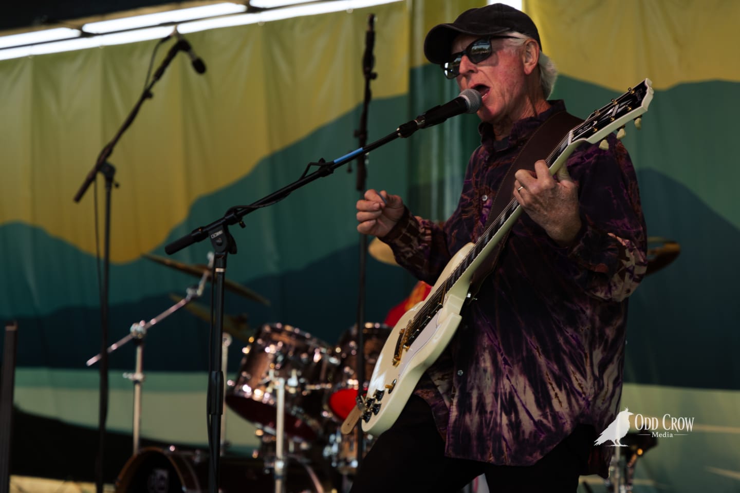 Older male guitarist singing into microphone on outdoor stage, holding white electric guitar, wearing cap and sunglasses.