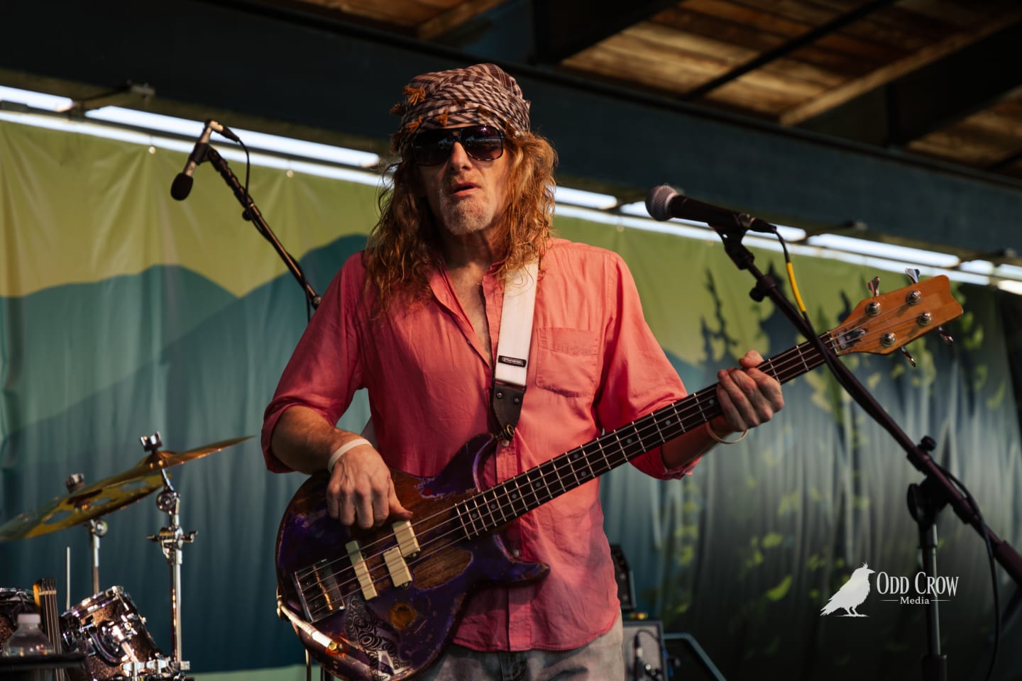 Redheaded bassist in pink shirt and bandana plays electric bass on an outdoor festival stage.