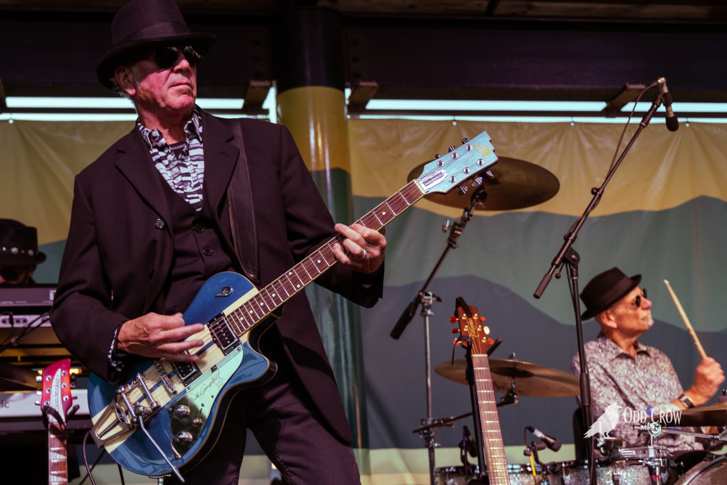 Petty Fever guitarist in black hat and suit playing a metallic blue Duesenberg guitar, drummer visible in the background