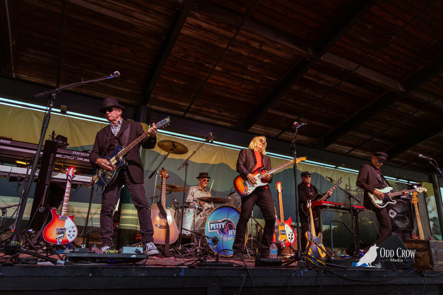 Full stage shot of Petty Fever with four guitarists plus drummer at Concerts on the Pier, Petty Fever bass drum logo centered in frame, mountain mural backdrop visible