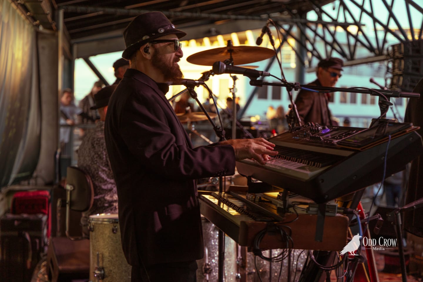Petty Fever keyboardist in black hat and dark jacket playing a Yamaha MODX M8 synthesizer, focused expression, stage lights behind him