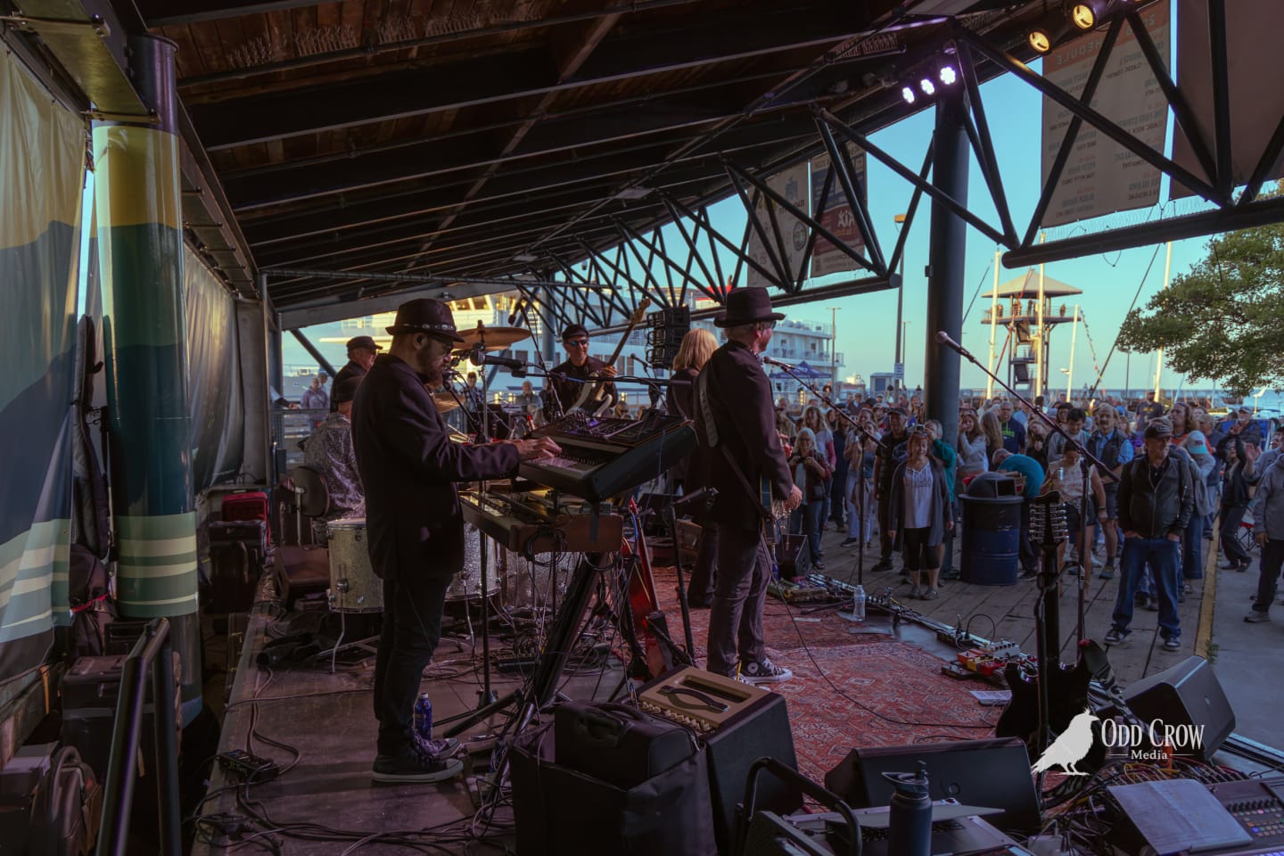 View from the back of the stage looking out at the large crowd gathered at the Port Angeles waterfront for Concerts on the Pier, evening light, ferry visible at the dock