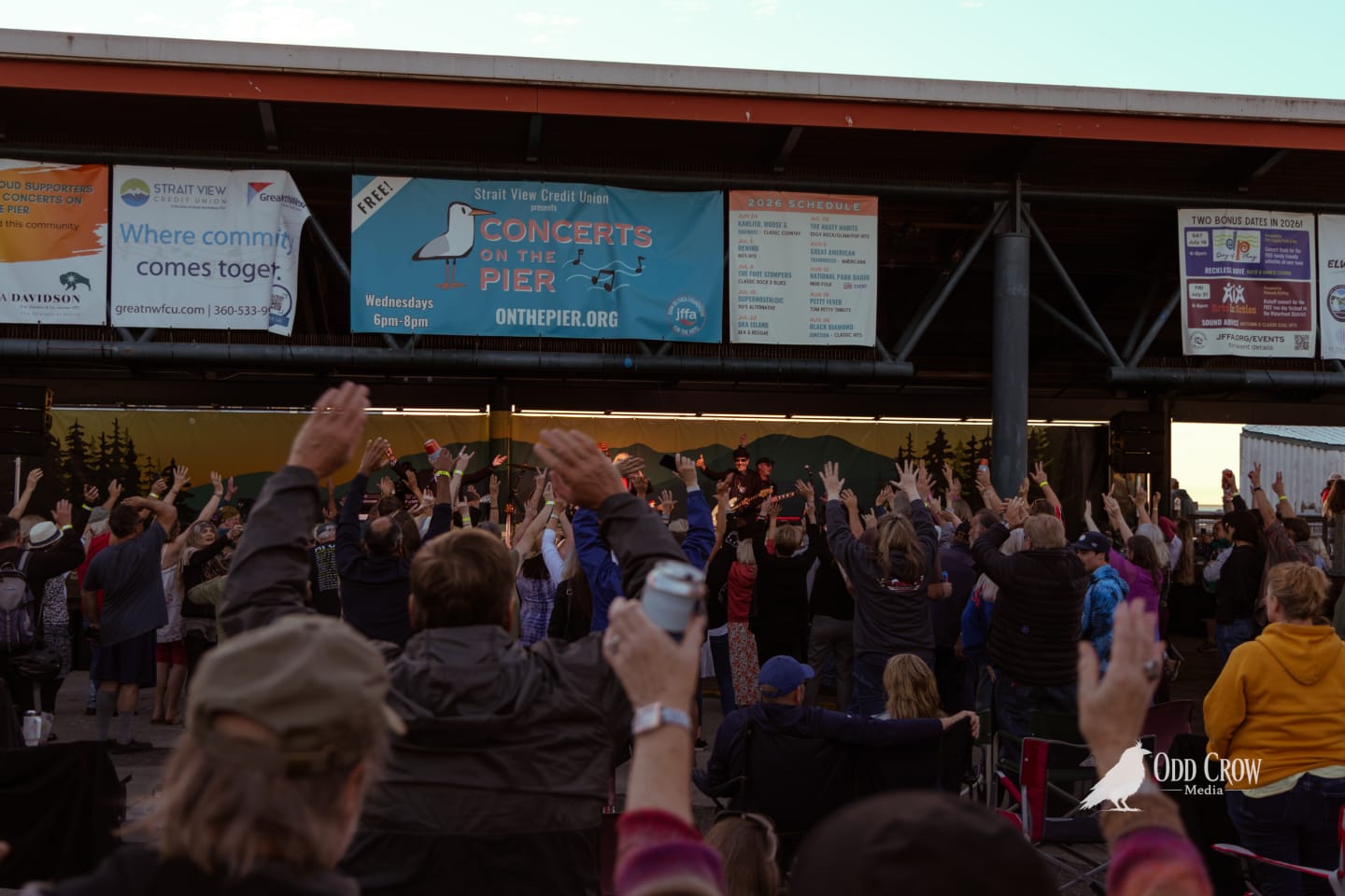 Crowd at Concerts on the Pier Port Angeles with hands raised during Petty Fever performance, banner reading Concerts on the Pier Wednesdays 6pm-8pm visible above