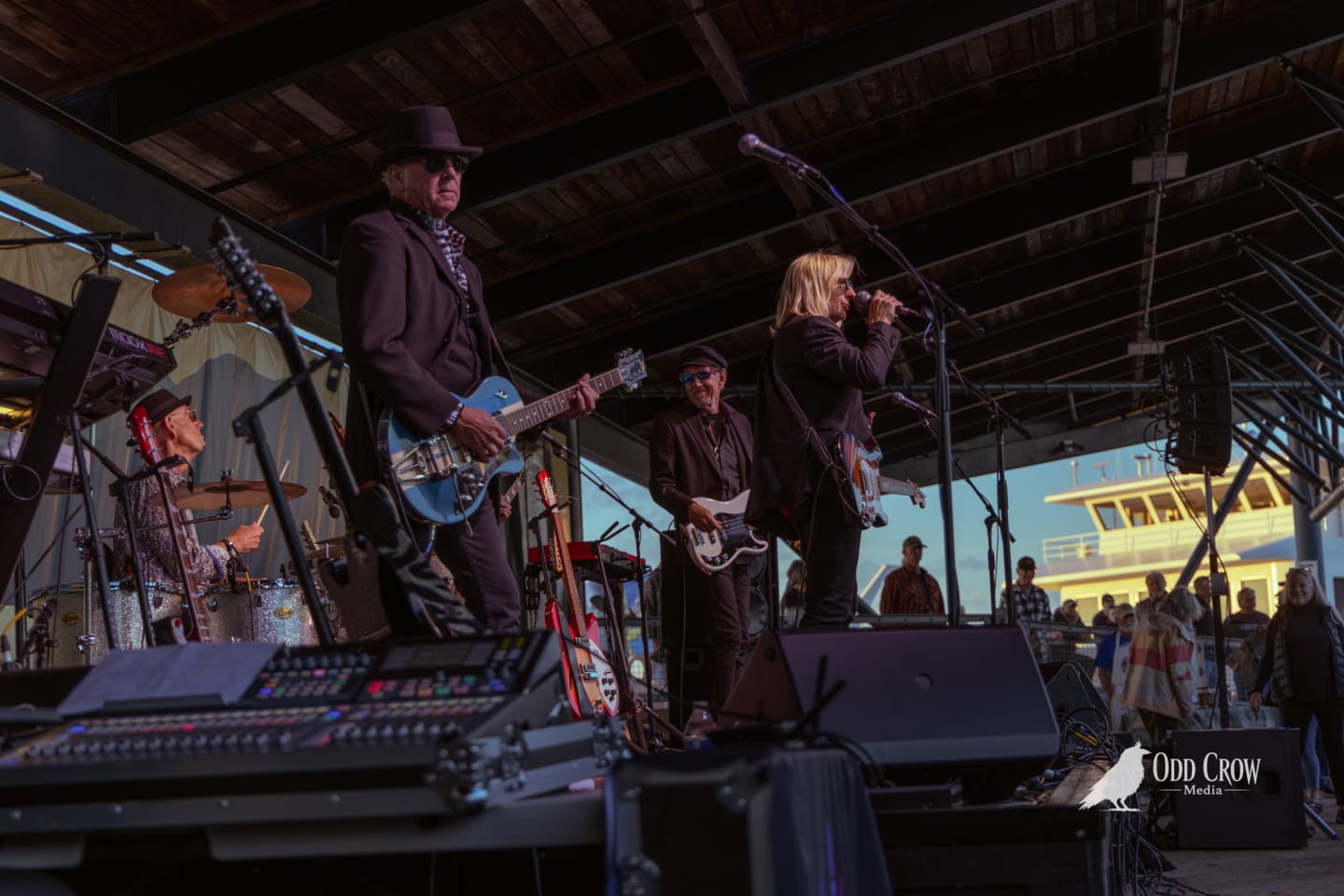 Petty Fever singer in red shirt and black blazer performing at the mic with two guitarists behind him, Port Angeles ferry terminal glowing in background at dusk