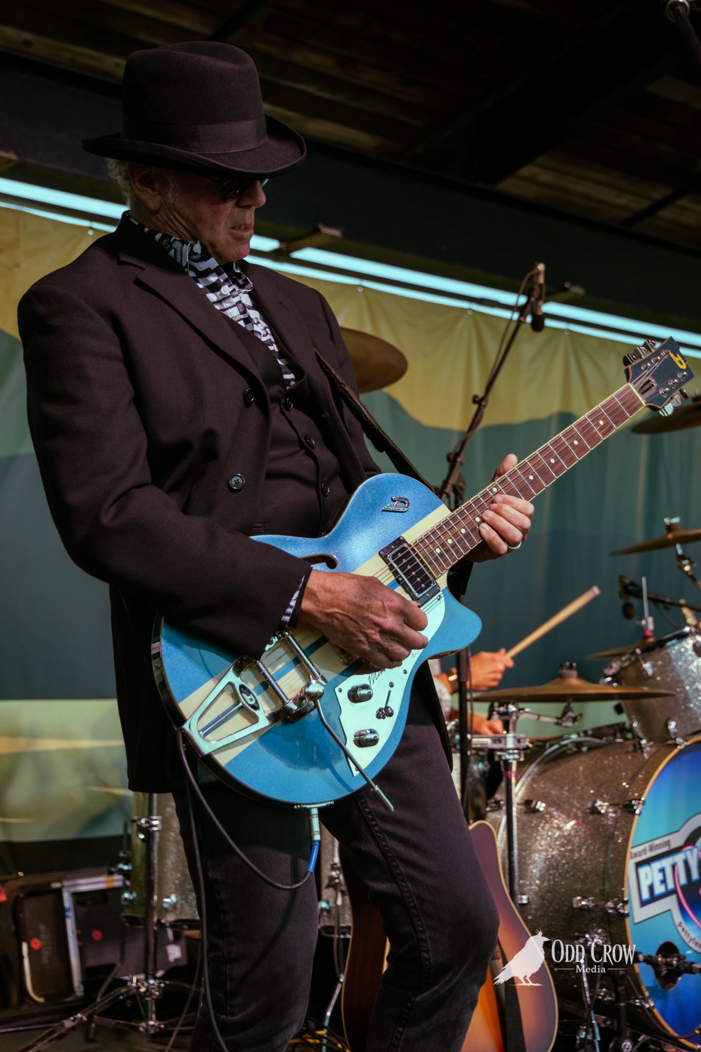 Petty Fever guitarist in black hat and zebra-print scarf playing a black 12-string electric guitar, multiple guitars on stands visible in the background