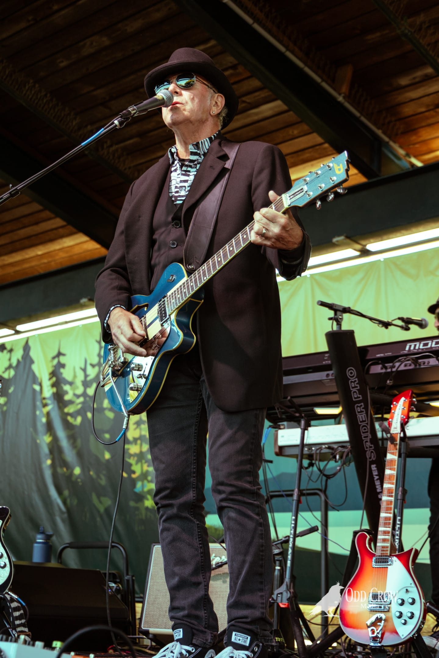 Petty Fever singer playing Takamine acoustic guitar, singing with eyes focused, drummer behind him in the background