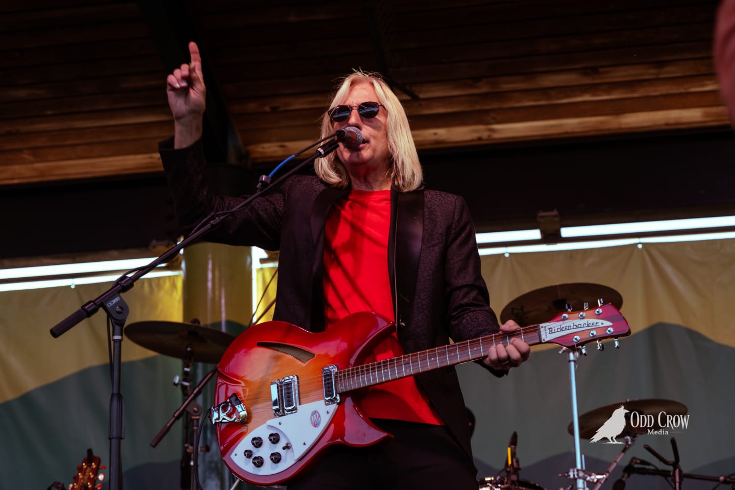 Petty Fever singer in red shirt and tall dark top hat performing at the mic with Rickenbacker guitar, stage lights flaring dramatically around him