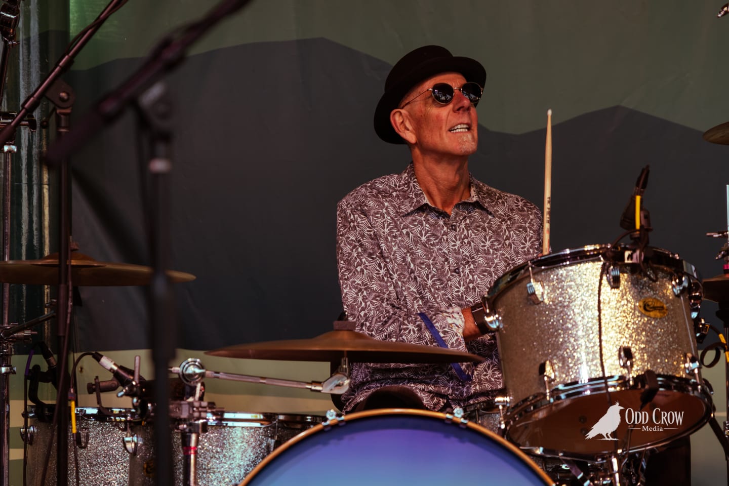 Petty Fever singer in top hat and red shirt playing Rickenbacker guitar, sun flaring through the open-air stage, bassist and drummer visible behind him