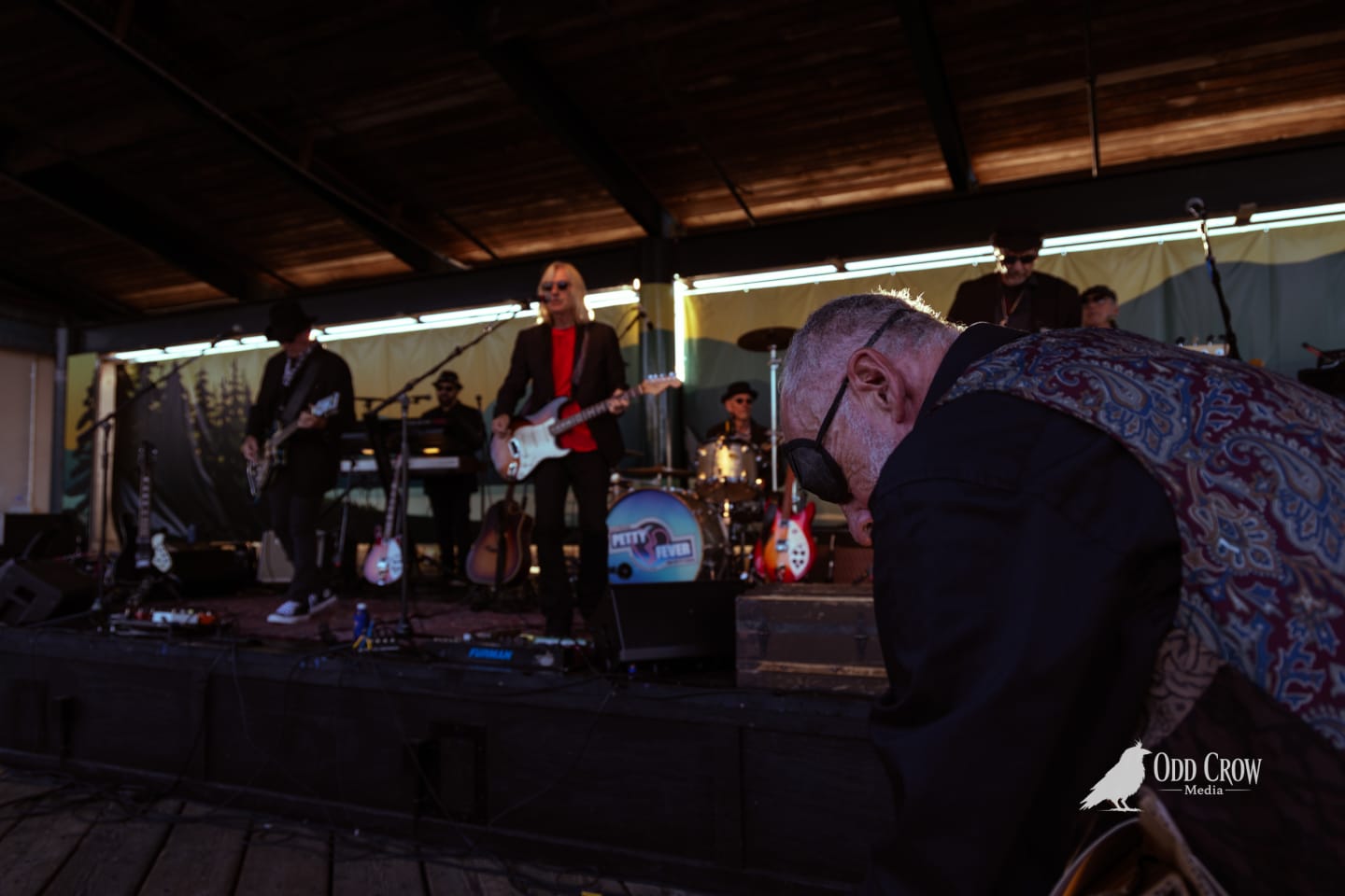 Artist in patterned vest bending over drawing board near the Petty Fever stage, band performing in background, stage lights illuminating the scene
