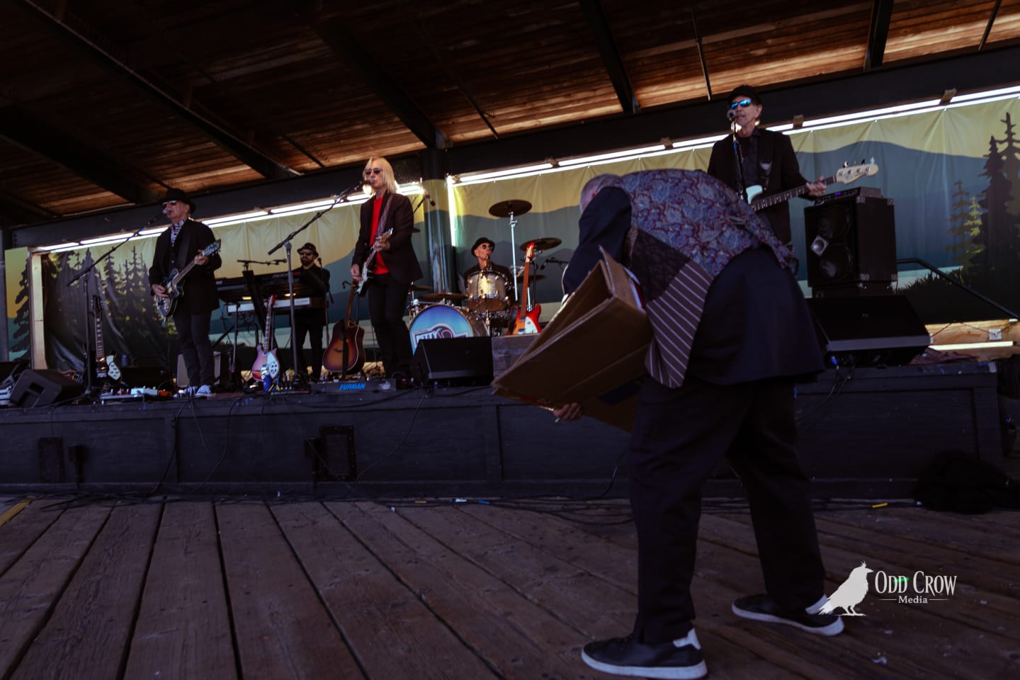 Artist in paisley vest holding a drawing board while Petty Fever performs on stage behind him, warm stage and evening light, lens flare streaking across the frame
