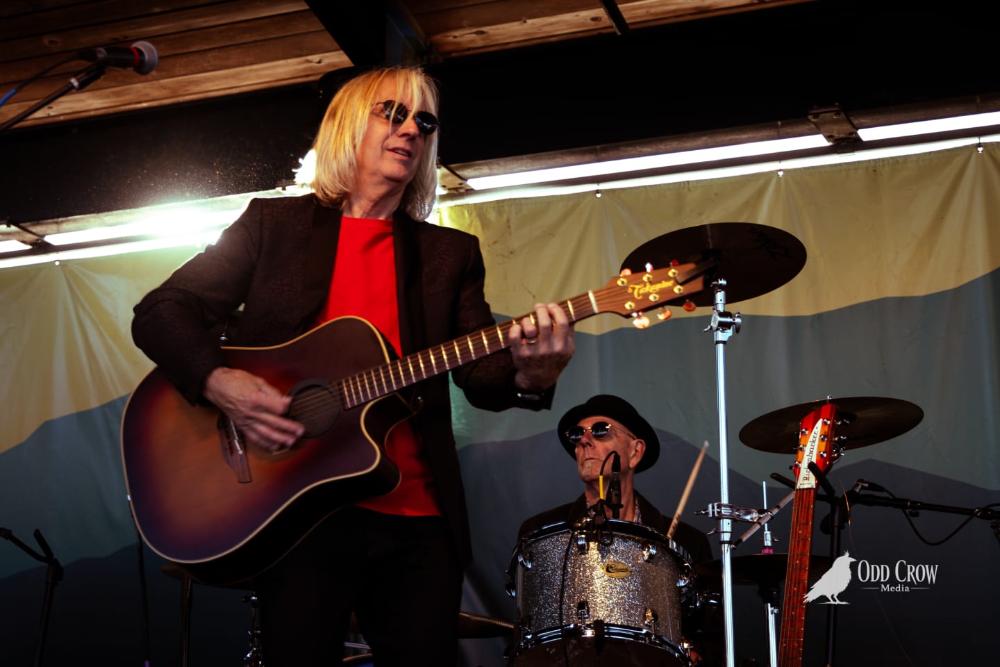 Petty Fever bassist singing at the mic in close-up, blue sunglasses catching the light, second guitarist visible smiling behind him