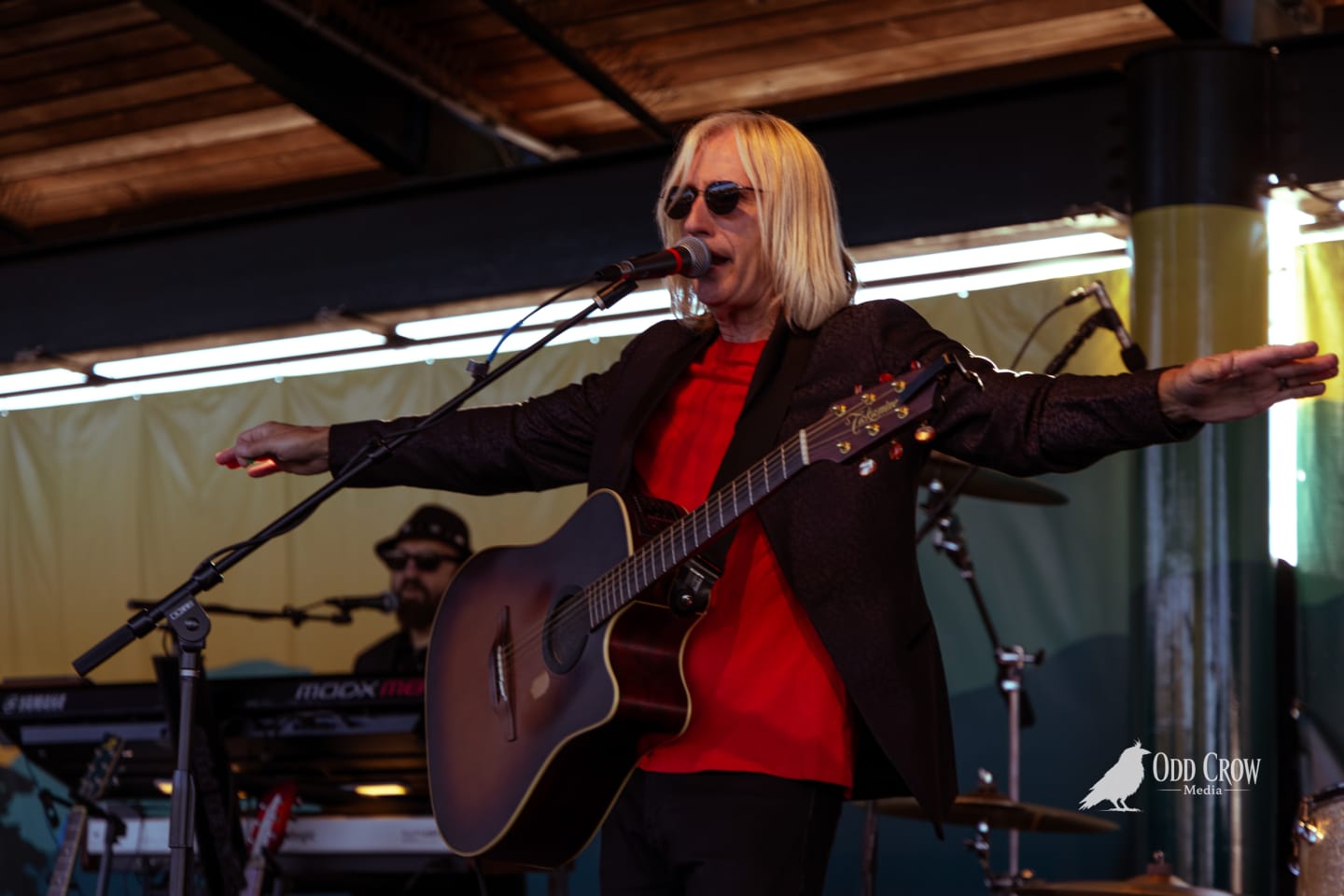 Petty Fever singer in red shirt and black blazer, Rickenbacker Fireglo guitar in hand, relaxed and confident, Petty Fever bass drum logo visible behind
