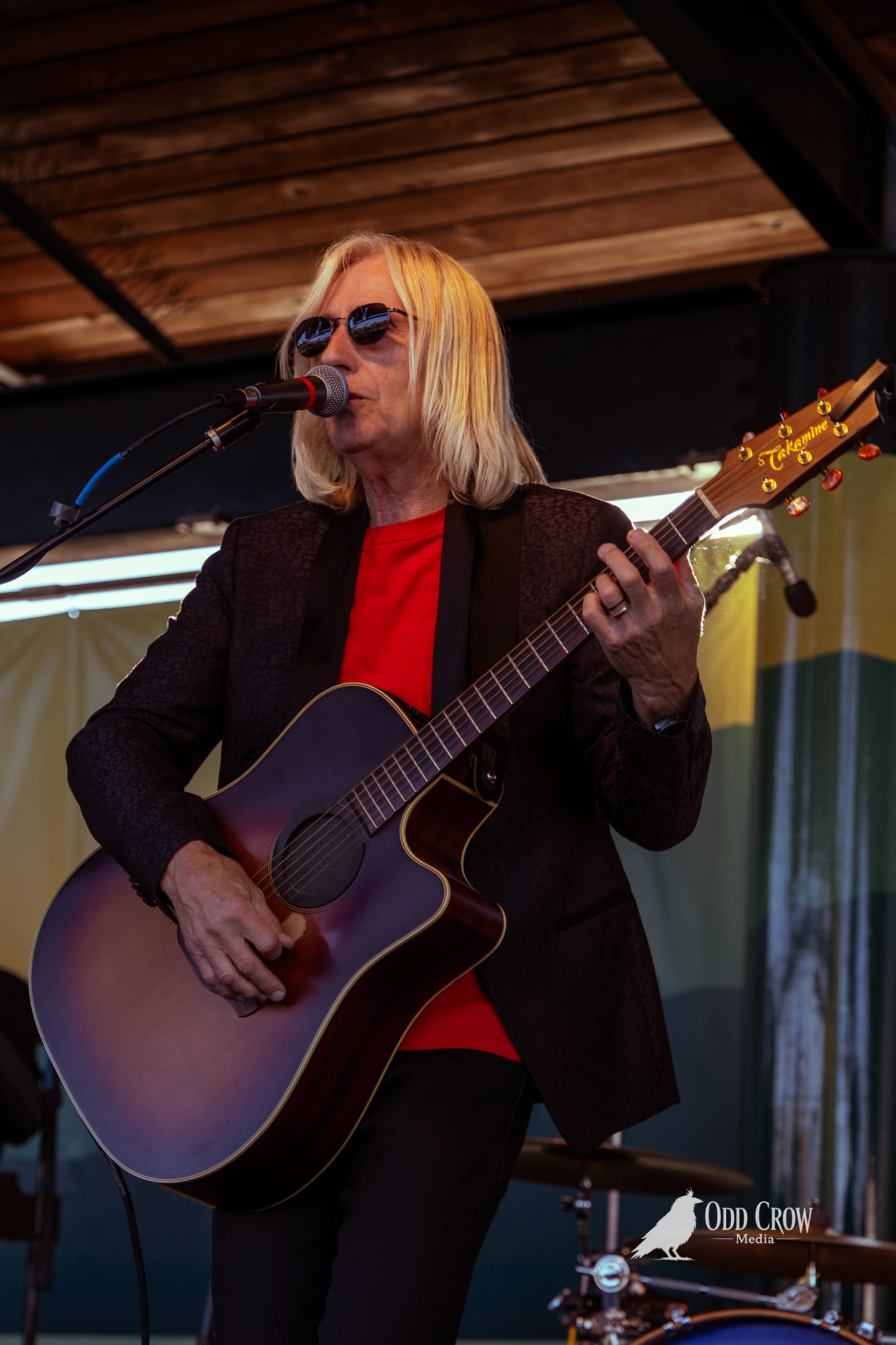 Petty Fever guitarist in black hat playing blue Duesenberg guitar and singing, sunlight streaming through the open-air stage from the side