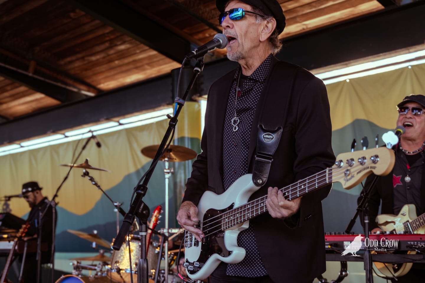 Petty Fever bassist in black cap with blue mirrored glasses looking directly at camera, slight smile, stage backdrop behind him