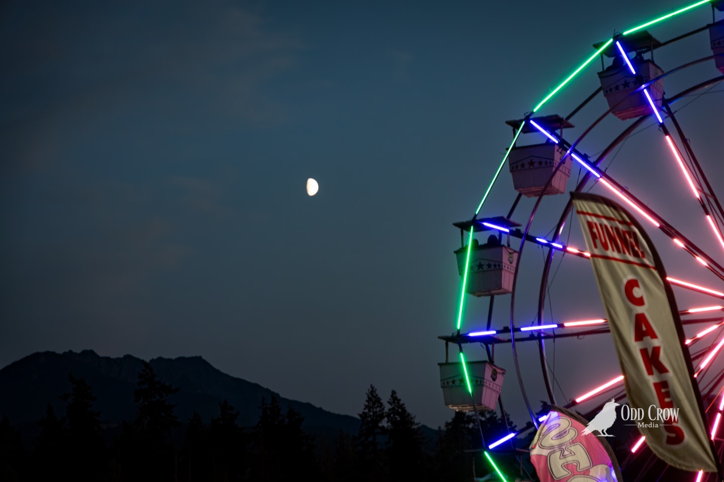Clallam County Fair Ferris wheel glowing with neon green, blue, and pink lights at dusk with a half moon rising over the dark silhouette of the mountains
