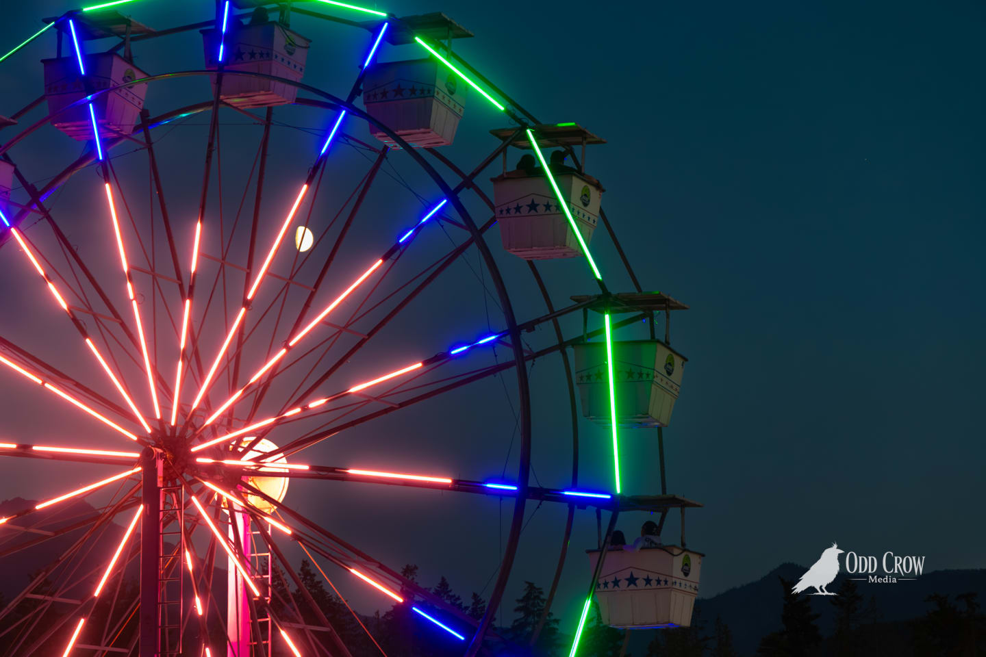 Long exposure of the illuminated Clallam County Fair Ferris wheel at night, neon spokes blurring against a dark sky with the moon visible through the wheel's center