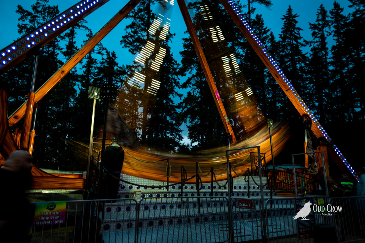 Long exposure of a swinging carnival ride at the Clallam County Fair, motion blur creating streaks of warm amber light against dark fir trees at blue hour