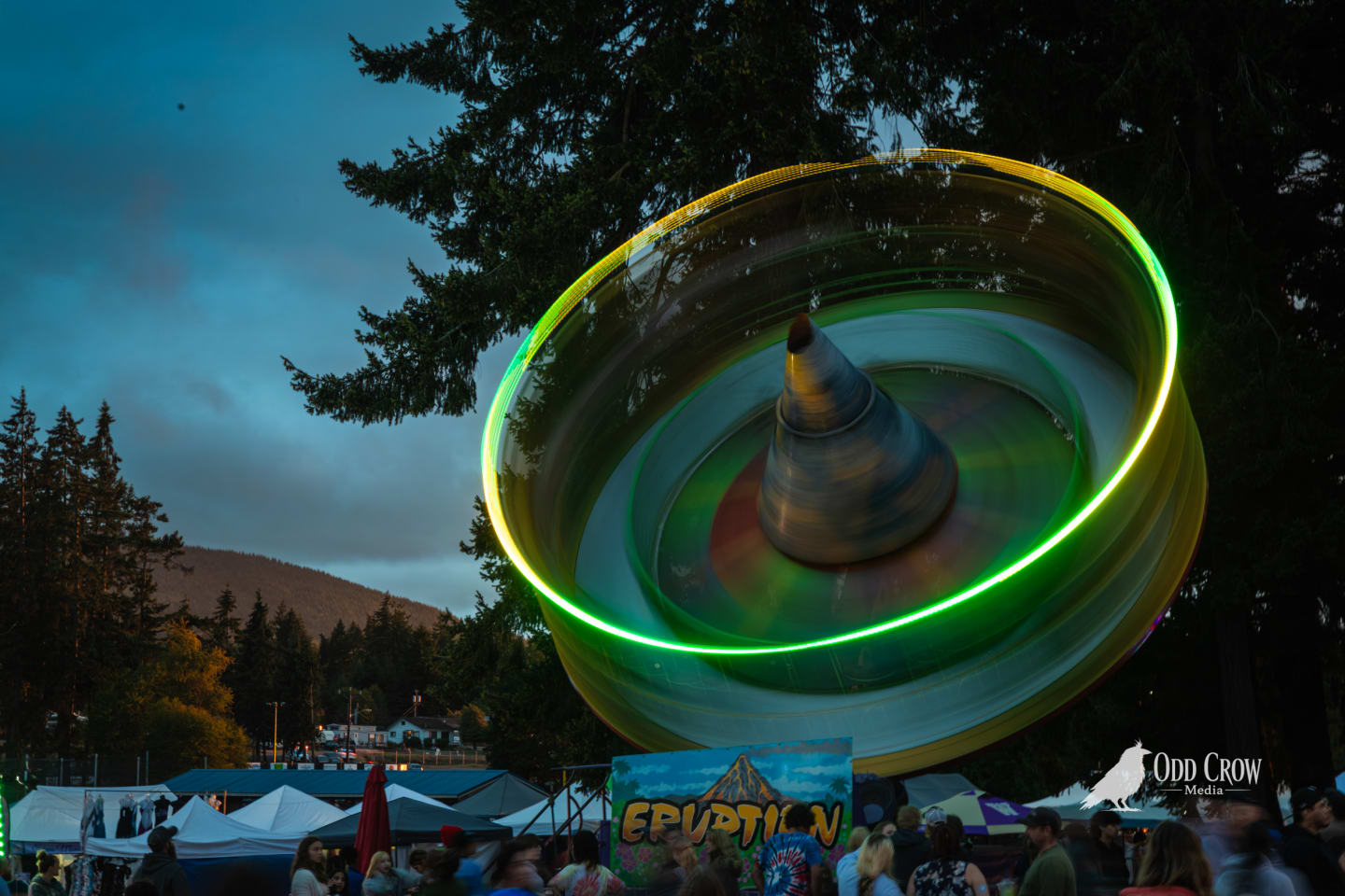 Long exposure of the Eruption spinning carnival ride at the Clallam County Fair, glowing yellow-green ring of light trails visible against dusk sky with crowd below