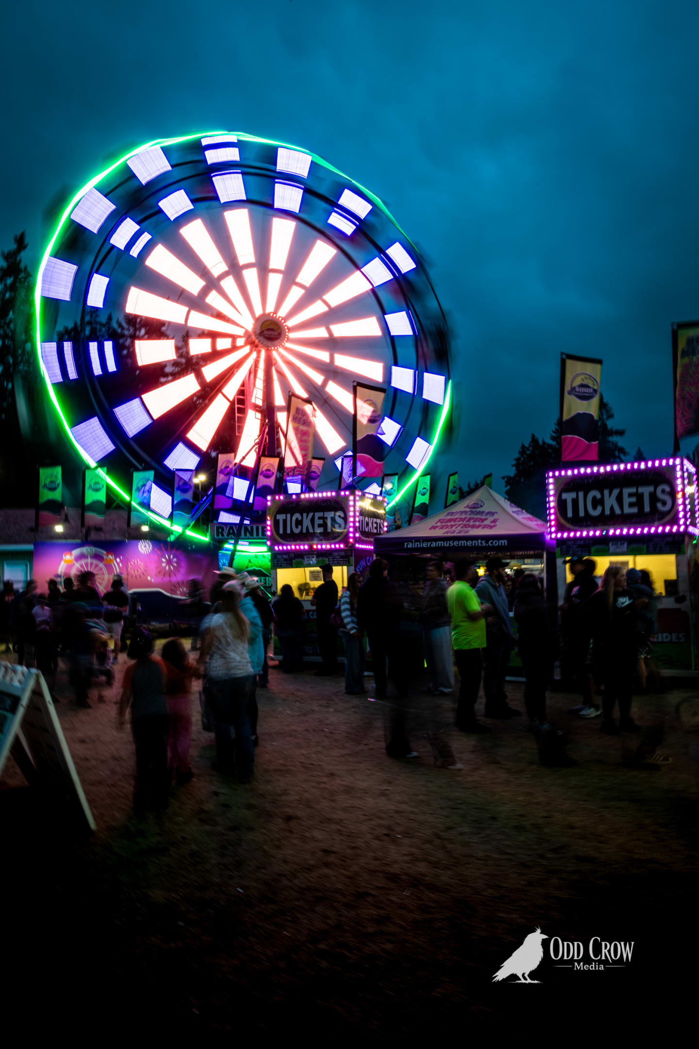 Long exposure of the Rainier Wheel Ferris wheel at the Clallam County Fair at night, spinning motion creating a glowing disc of white, green, and red light against a dark blue sky