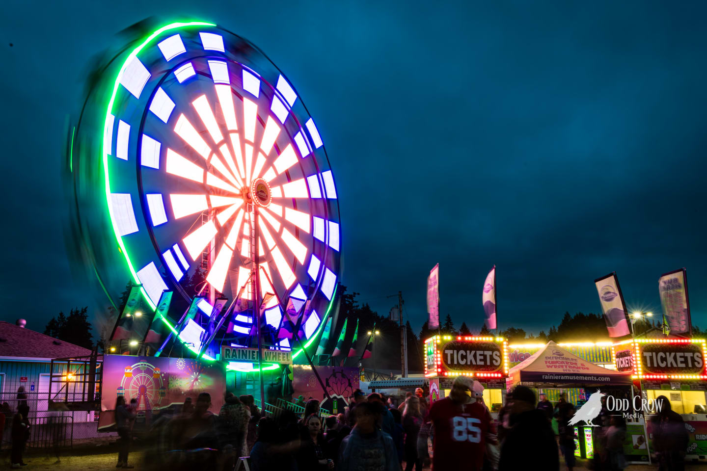 Wide nighttime view of the Rainier Wheel Ferris wheel spinning at the Clallam County Fair with crowd walking the midway below, ticket booths glowing in the background