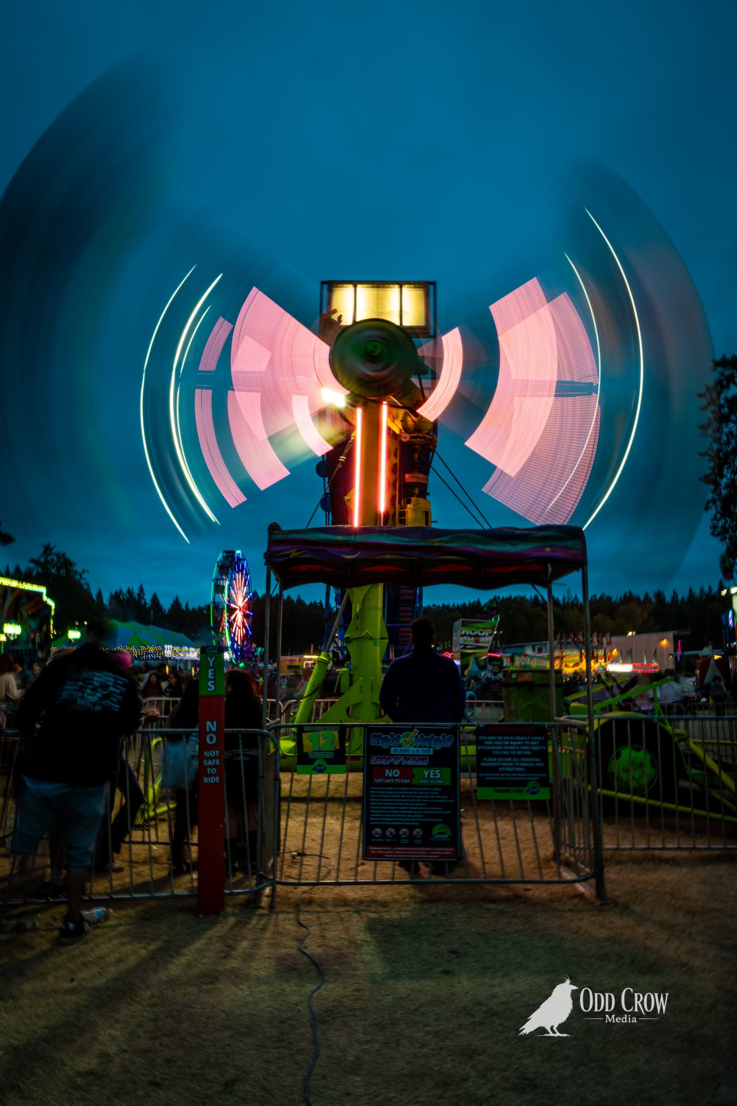 Second long-exposure shot of the spinning gyroscope ride at the Clallam County Fair, symmetrical arcs of light emanating outward against a blue dusk sky
