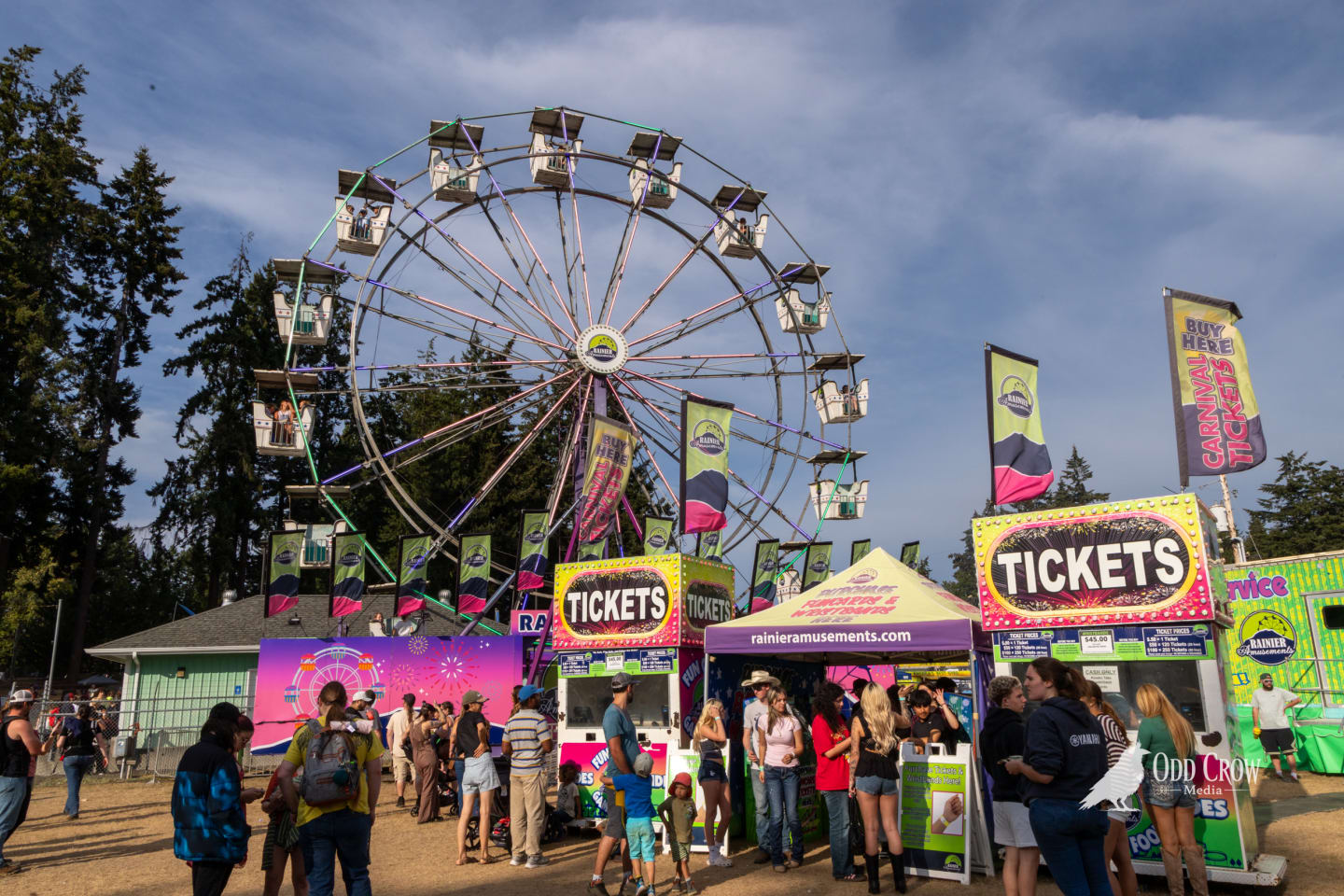 Daytime view of the Clallam County Fair midway with the Ferris wheel against a partly cloudy blue sky, crowds lining up at Rainier Amusements ticket booths