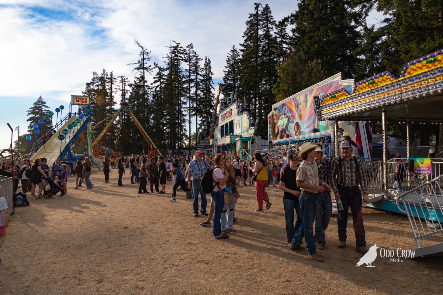 Clallam County Fair carnival midway in late afternoon light, crowds walking between rides including a super slide and colorful attraction buildings under tall fir trees