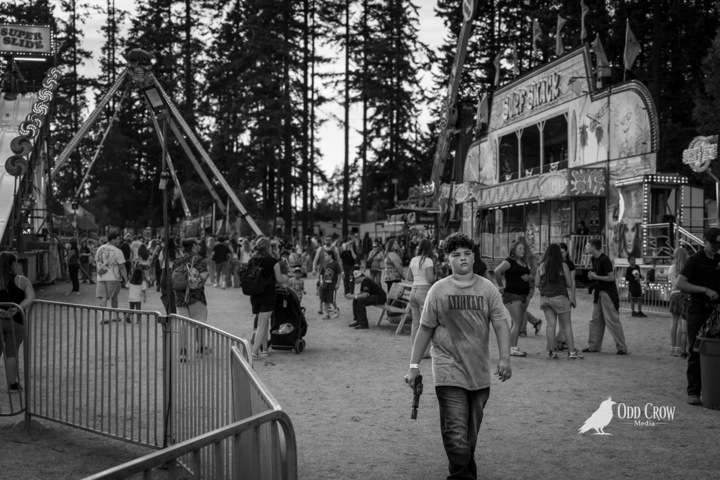 Black and white photo of a teenager in a Nirvana t-shirt walking the Clallam County Fair midway at dusk, fairground rides and crowds visible in the background