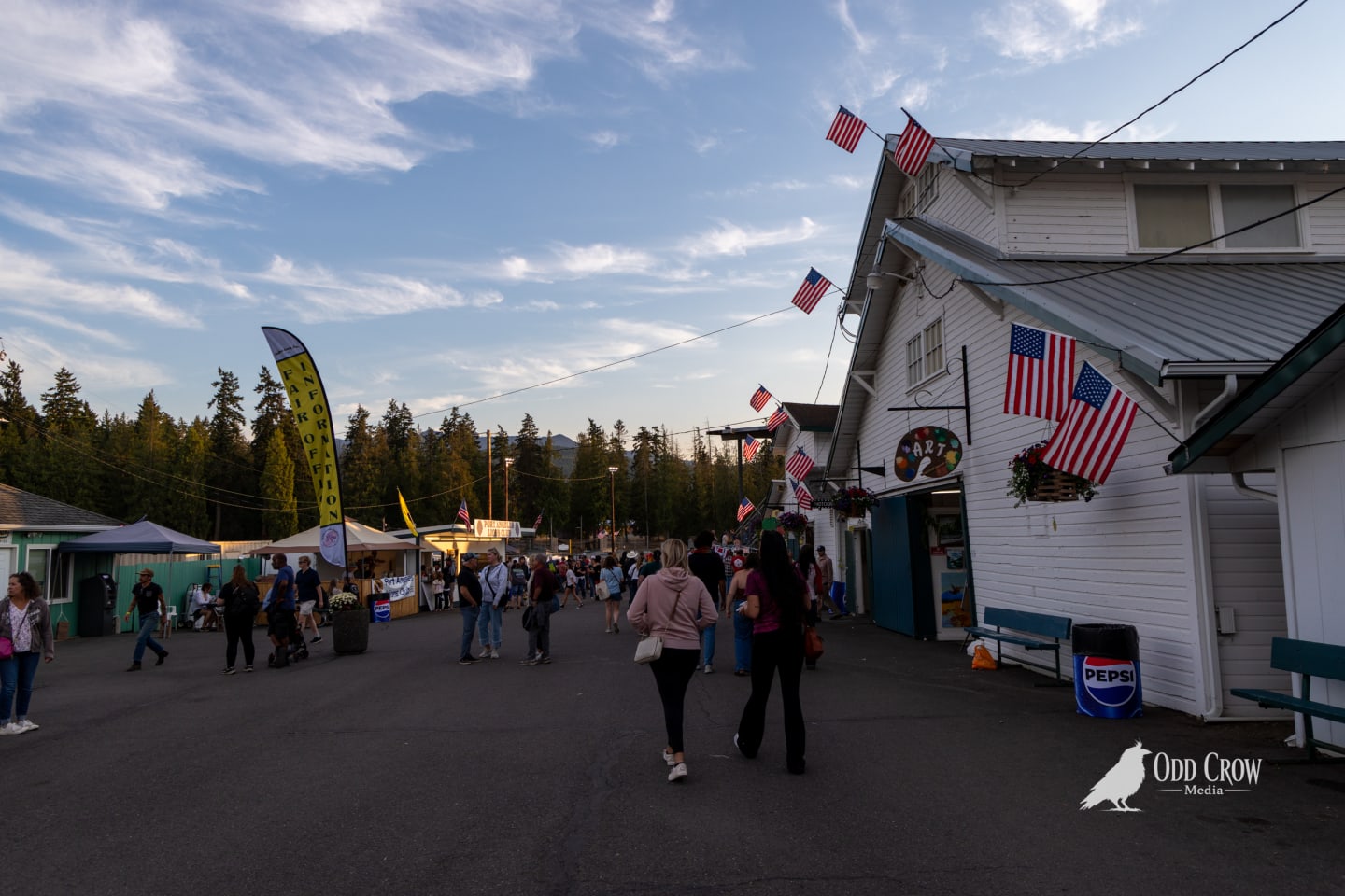 Wide view of the Clallam County Fairgrounds main grounds at golden hour, white exhibition buildings with American flags, attendees walking between vendor tents and displays