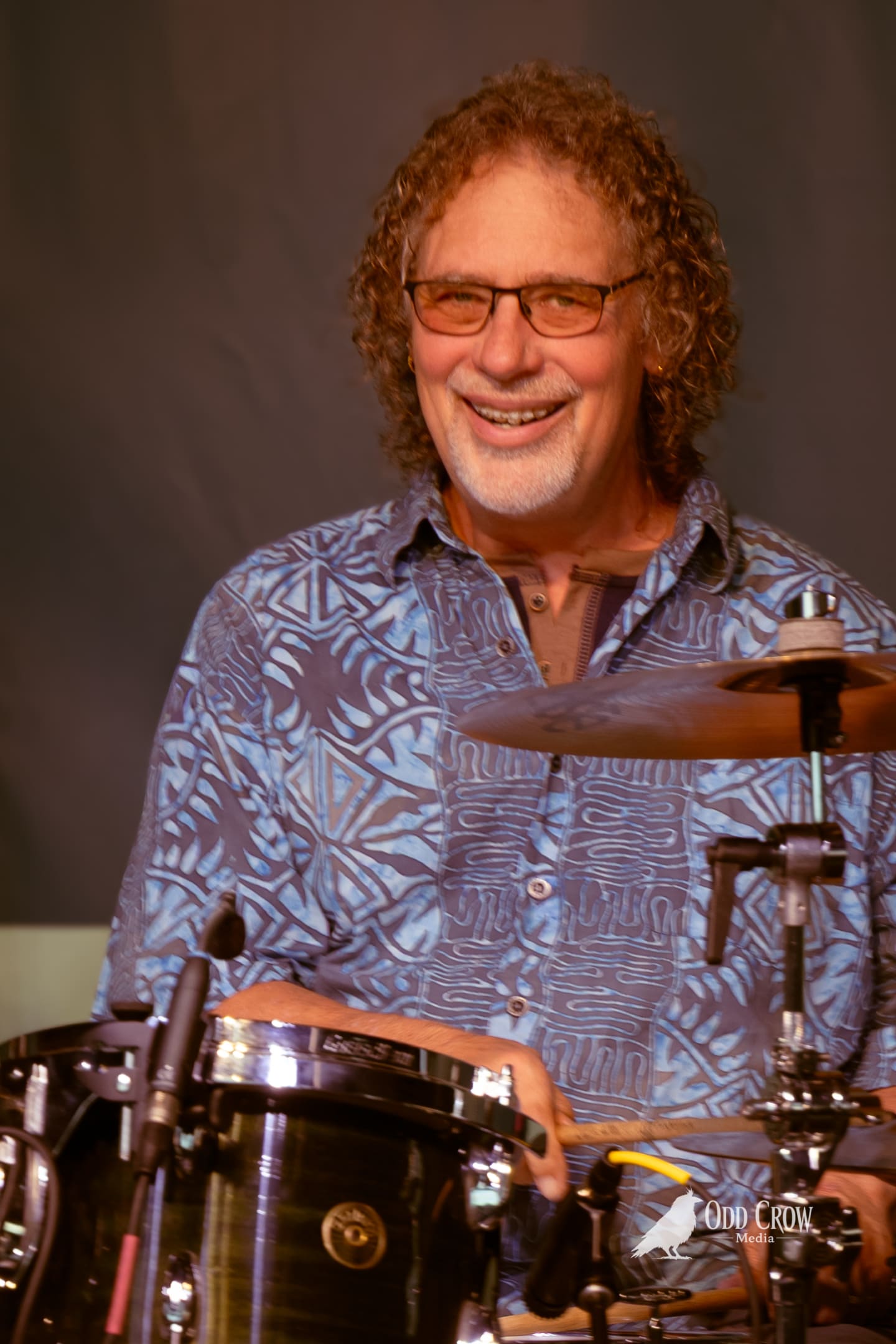 Smiling drummer with curly hair and glasses plays kit on outdoor stage at pier concert.